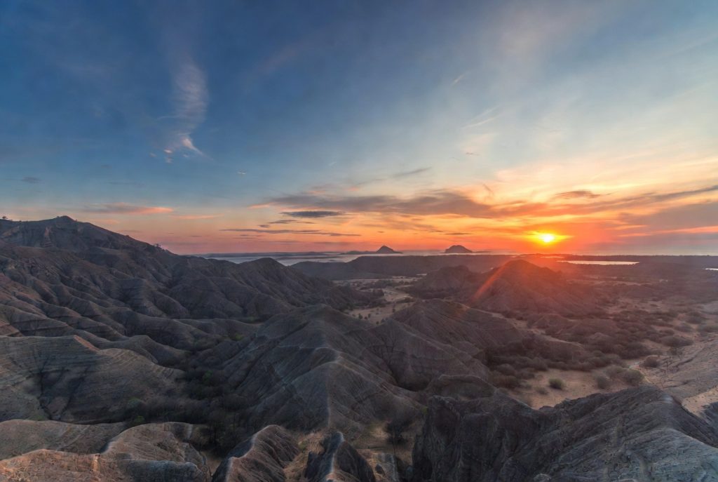Sunrise dramatis di puncak Pulau Padar dengan siluet bukit savana, laut biru turquoise, dan langit jingga keemasan – momen ikonik travel NTT yang memukau hati